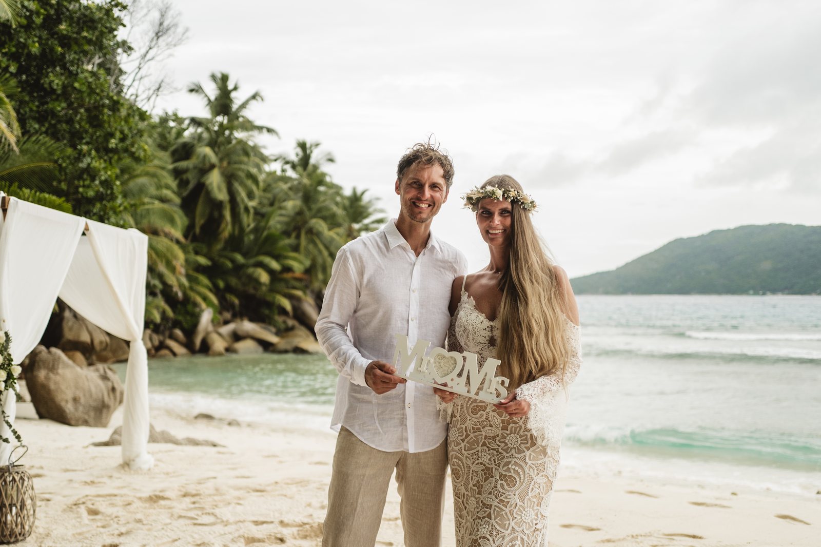 A couple on the sand in front of white drapes and greenery