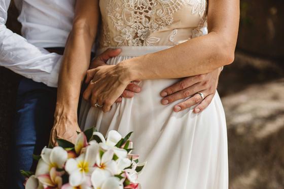Hands folded at her waist, rings showing, frangipani bouquet below