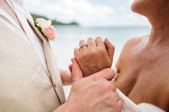 Clasped hands with a rose boutonniere, the beach soft behind