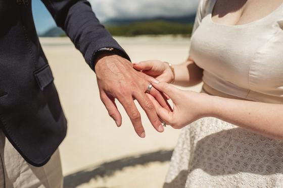 The bride sliding the ring onto the groom's hand, beach behind